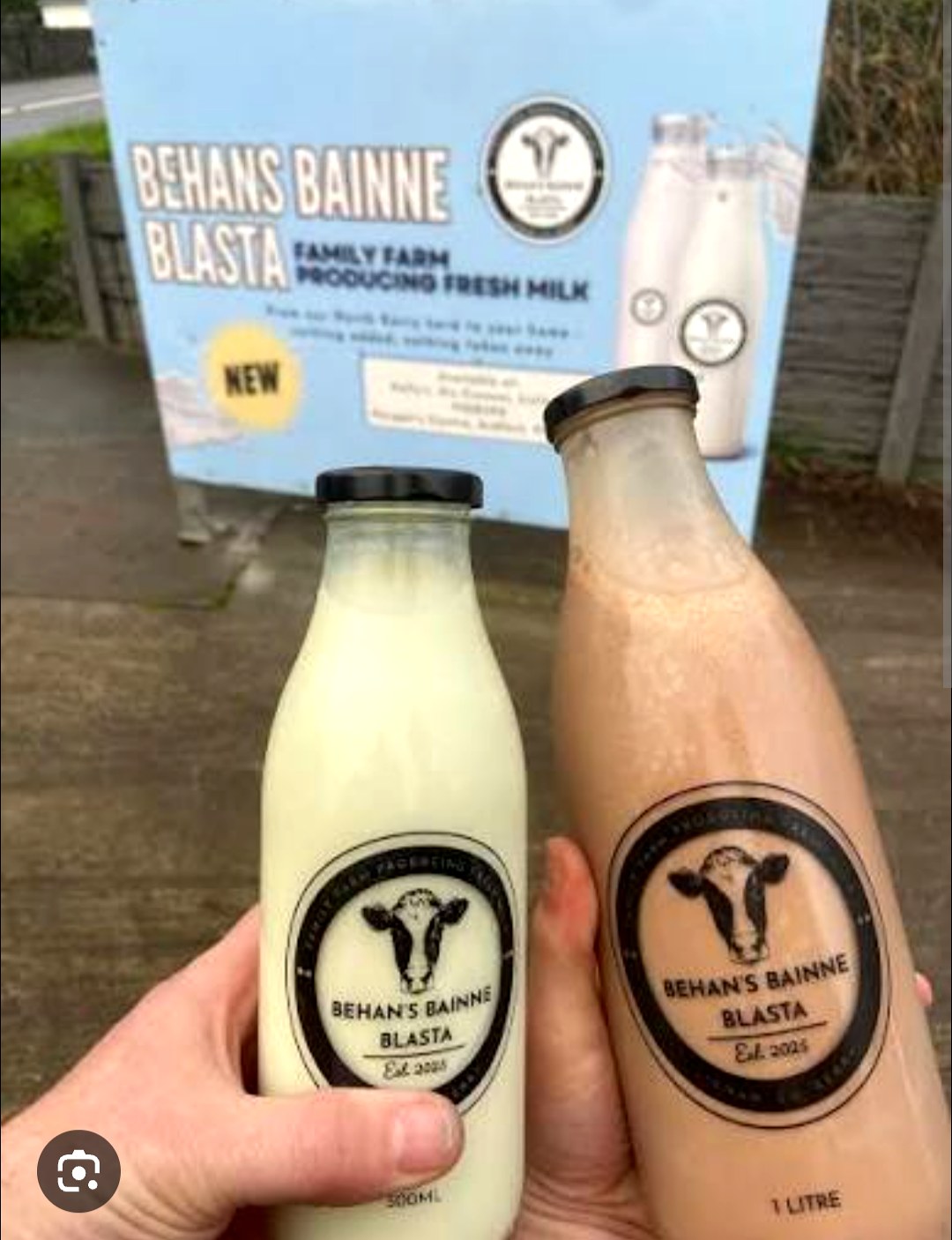 A 500ml fresh milk bottle and a 1 litre chocolate bottle held in hand in front of the Behan's Bainne Blasta farm sign